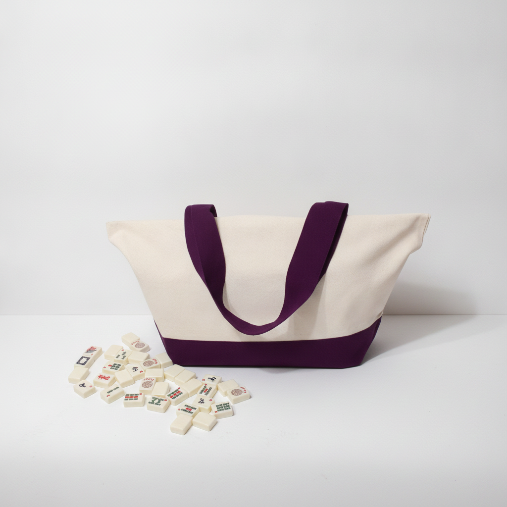 A canvas tote bag with a cream body and dark purple handles/bottom trim, shown on a white studio surface with a small scattered group of white game tiles in front.