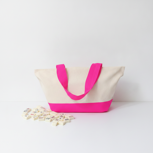 A canvas tote bag with a natural beige body, bright hot pink handles, and a matching hot pink bottom band, displayed on a white surface with small decorative blocks in the foreground.