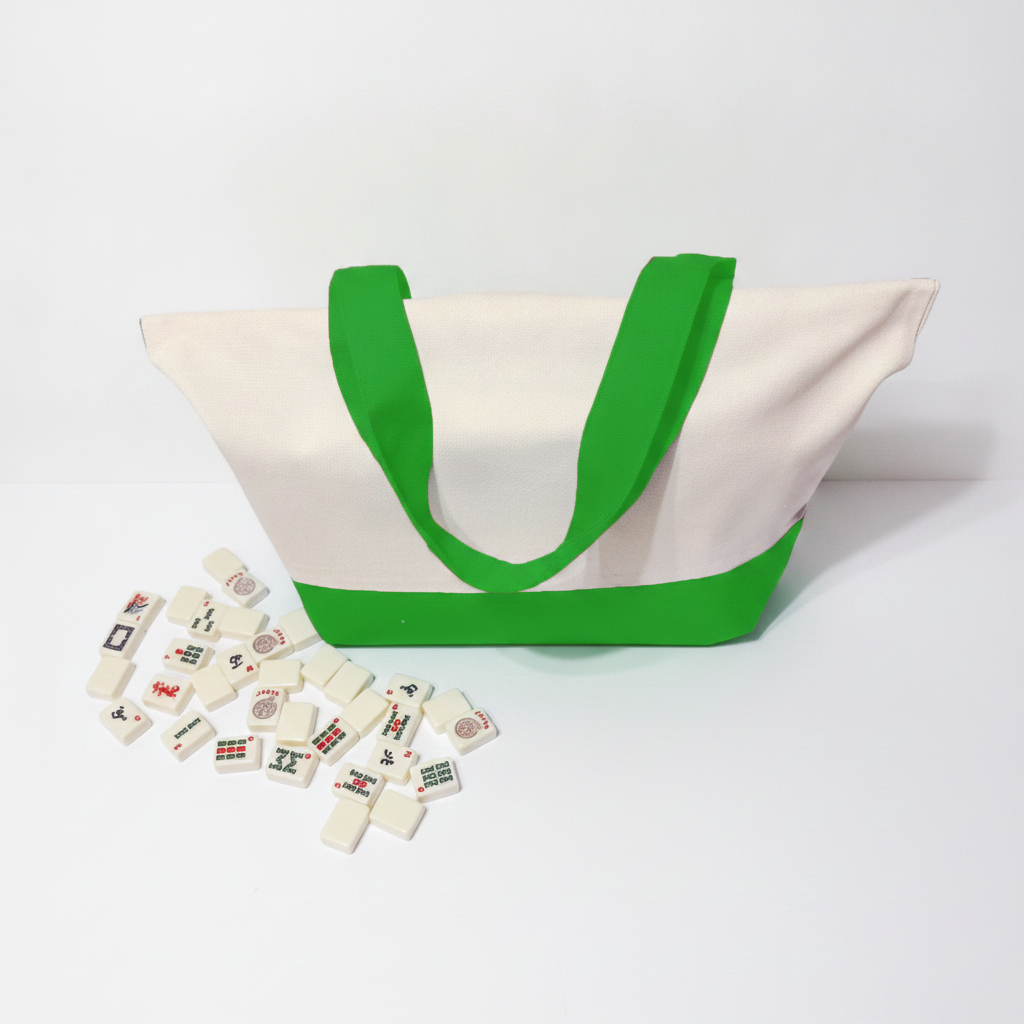 A canvas tote bag with a cream-colored body and bright green handles, with a few mahjong tiles scattered in front on a white studio background.