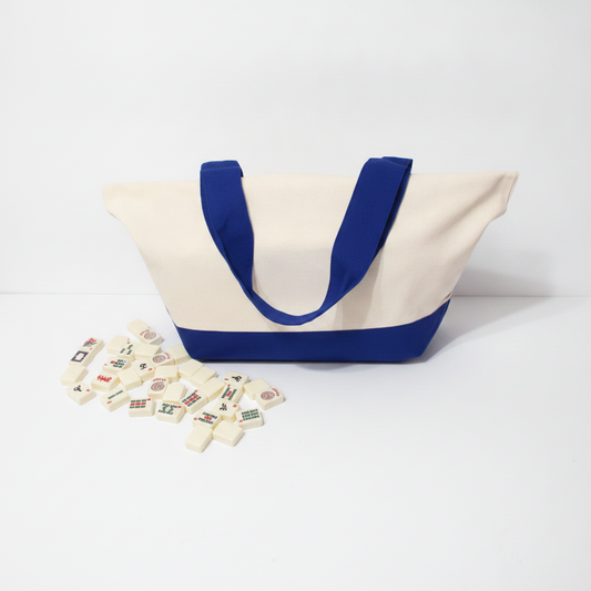 A canvas tote bag with a cream body and royal blue handles, bottom trim, and interior lining, photographed on a clean white studio background. A small scattered cluster of rectangular game pieces lies in front of the bag.