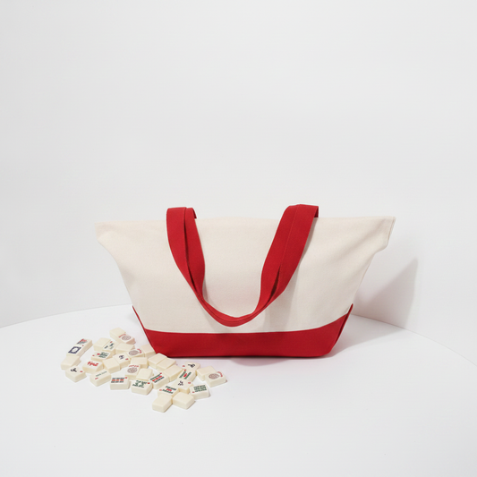 A canvas tote bag with a cream-colored body and red handles, bottom trim, and base, displayed on a white surface with a few small decorative tiles scattered in front.