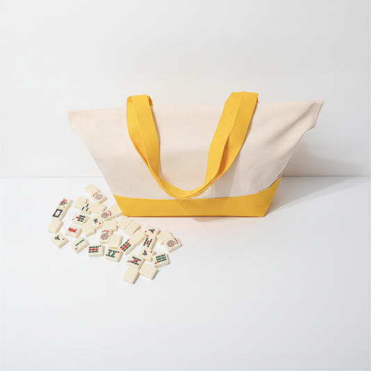 A canvas tote bag with a cream body, mustard yellow handles, and a mustard yellow bottom band, shown on a white studio background with small game pieces scattered in front.