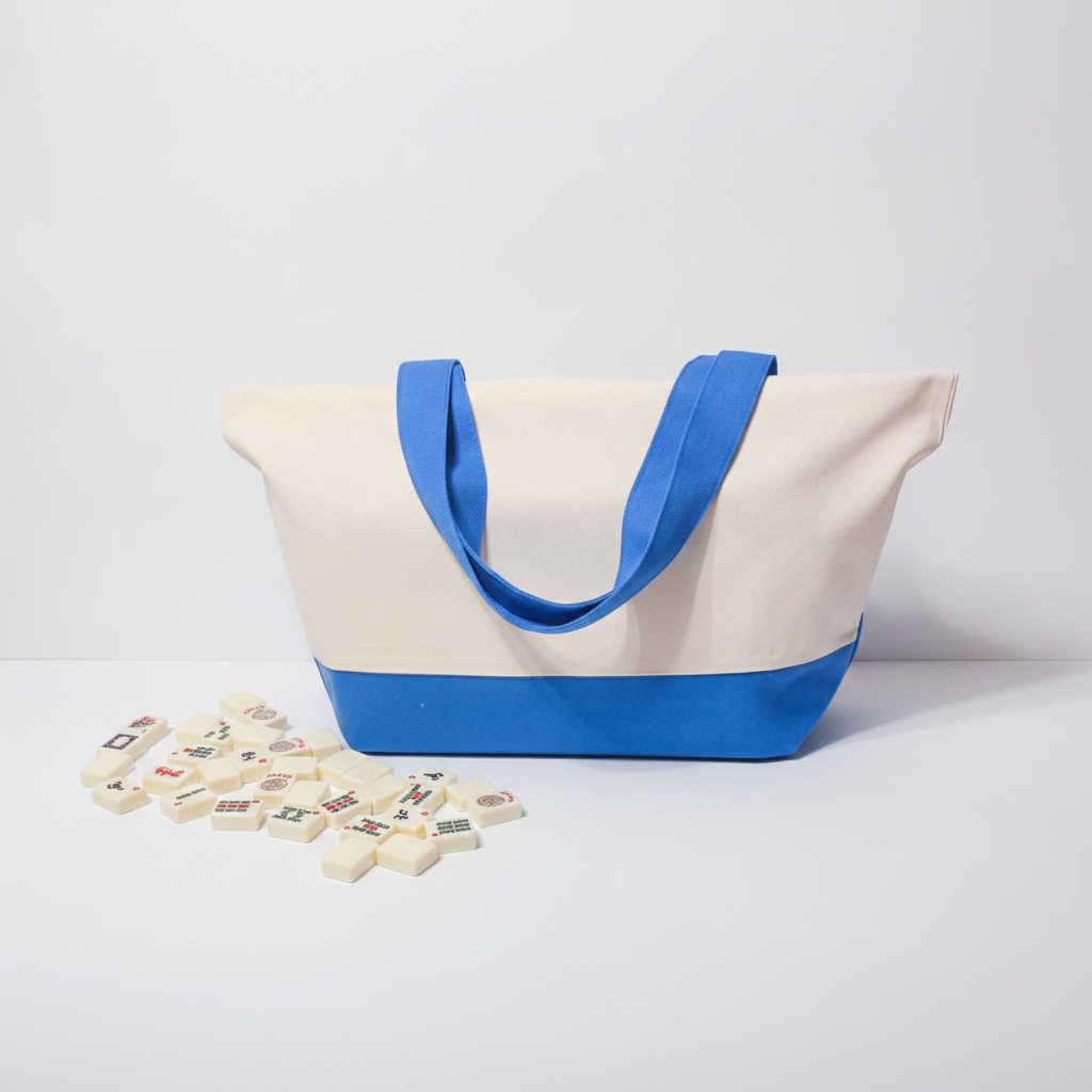 A canvas tote bag with a cream body, blue handles, and a blue bottom band, photographed on a clean white studio surface. A small cluster of rectangular decorative tiles lies in front of the bag.