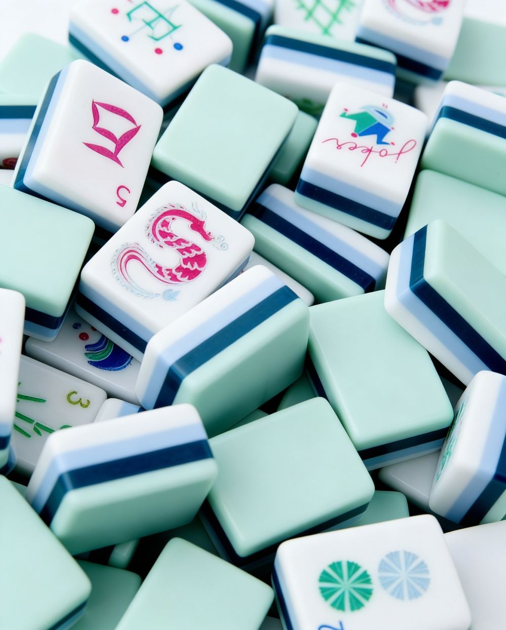 A close-up pile of  mahjong tiles in mint green and white with blue stripes and colorful Chinese characters and symbols.